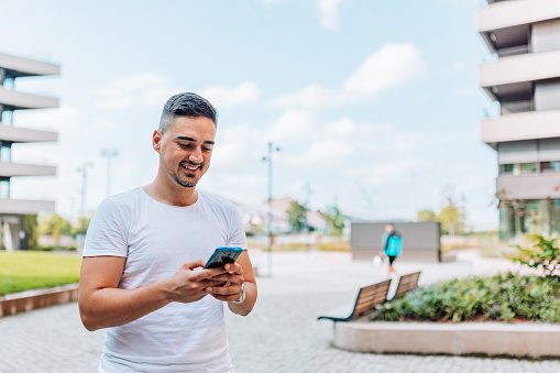 A young Caucasian man in a white shirt is looking at his phone with a smile on his face.