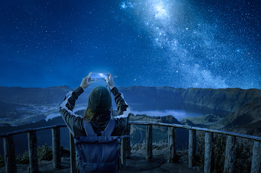Woman with a smartphone taking a photo of a beautiful night sky. Composed photo without a real night sky. Base photo taken near Sete Cidades on São Miguel Island, Azores.