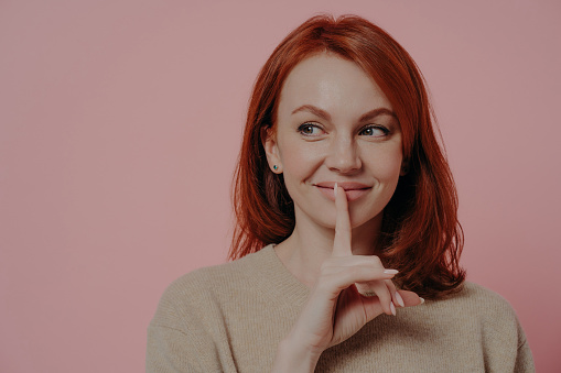 Close up of beautiful young smiling ginger woman putting finger on lips, making hush sign and looking aside at copy space, asking to be quiet, showing silence gesture while standing isolated on pink