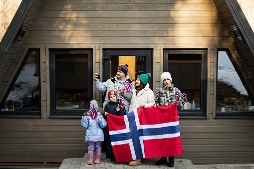 Portrait of family with kids outside cabin house holding Norway flags and making selfie on phone. Scandinavian culture, norwegian people.