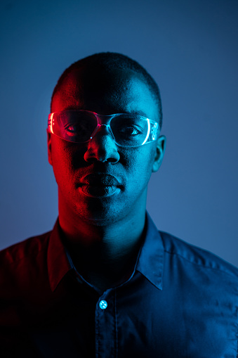 Headshort of an African-American man wearing glasses and a dark shirt while looking towards the camera, lights are reflected in the glasses against a dark background