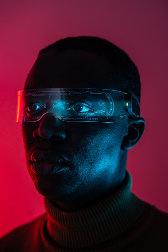 A close up of an African-American man, against a dark purple background but with light reflecting across his warp-around clear glasses and eyes
