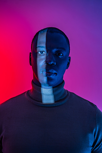 An African-American man with a vertical band of white light down one side of his face, wearing a turtleneck sweater against a red to purple background