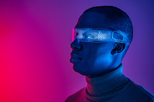 A side view headshot of an African-American man wearing wrap-around smart glasses and a rolled neck sweater against a gradient purple background