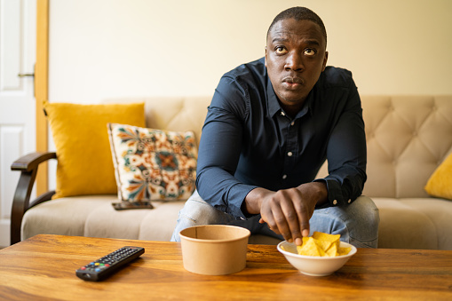 An Anglo-Caribbean man is concentrating on watching an unseen TV as he also reaches for a potato chip in a bowl on the coffee table in a modern living room