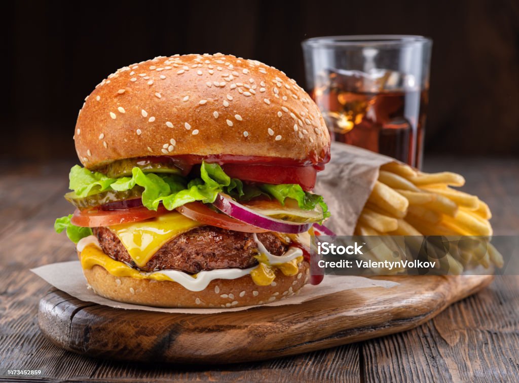 Tasty cheeseburger, glass of cola and french fries on wooden tray close-up. Tasty cheeseburger or hamburger, glass of cola and french fries on wooden tray close-up. Burger Stock Photo