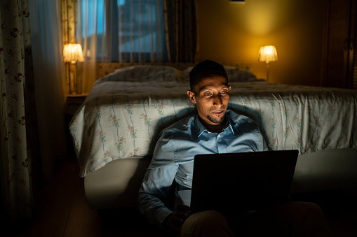 An African-American man is leaning against the end of his bed sitting on the floor while checking his laptop late at night