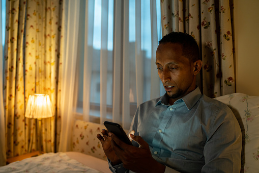 Close up of an African-American man holding a mobile phone in his hands while propped up against some pillows on his bed