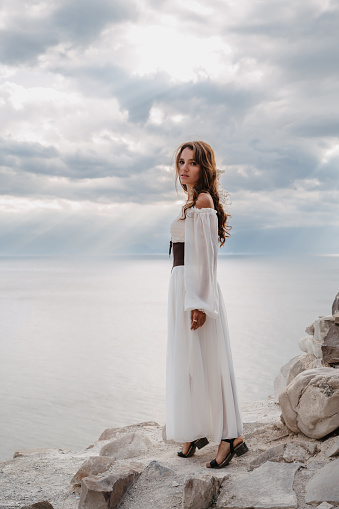 A young woman in a white dress stands barefoot on a cliff face in full height. The dress flutters in the wind. A sacred glow breaks through the clouds. The girl looks like an angel.