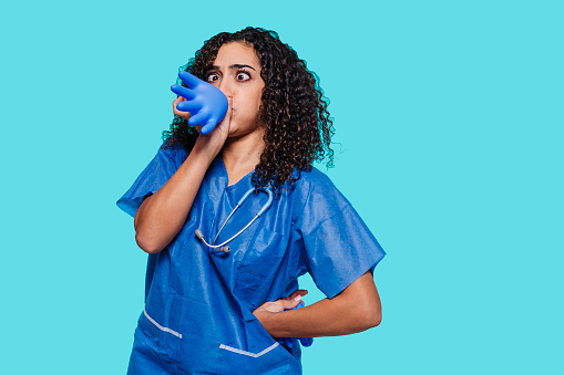 Young arabic surgeon woman inflates a plastic latex glove isolated over blue background. Funny facial expression, children pediatrician, nurse, health care concept.