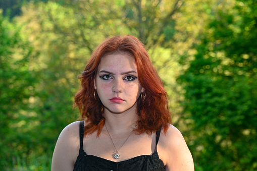 Portrait of a young pretty red-haired girl from the front, looks serious, in green nature
