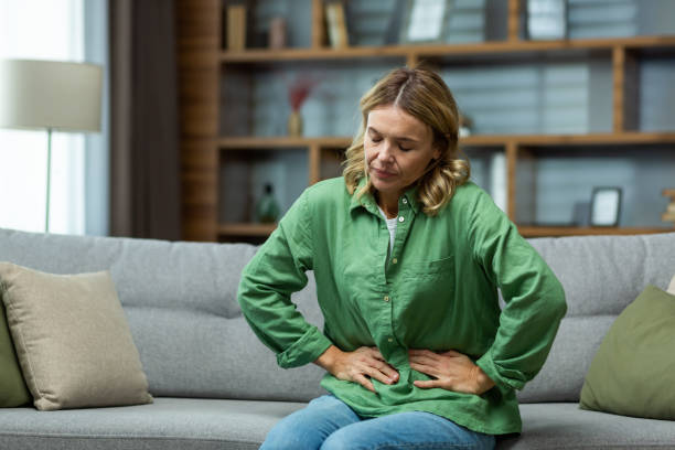 uma mulher idosa está sentada no sofá de casa, segurando o estômago com as mãos. sente a dor dos órgãos internos, indigestão, envenenamento, dor menstrual - intoxicação de substâncias - fotografias e filmes do acervo