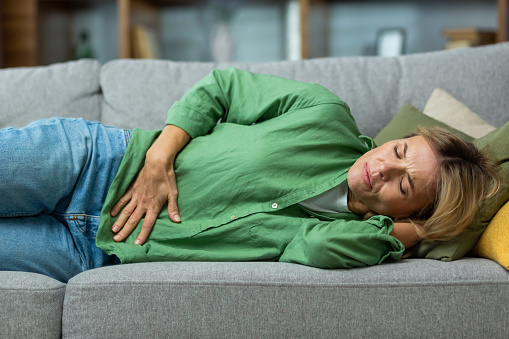 Mujer madura sola en casa enferma, cogida de las manos a un lado del estómago con un espasmo severo acostada en el sofá de la sala de estar photo