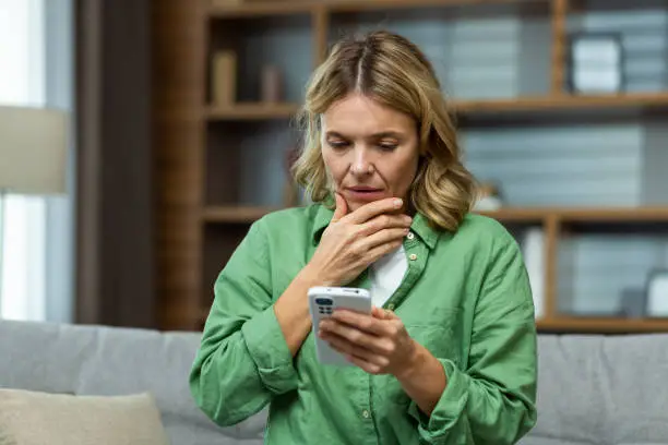 Photo of Close-up photo. Worried senior woman mother sitting on sofa at home and holding phone. Worries about children, writes and sends messages, calls, searches, waits at home