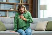 Senior beautiful woman sitting sick on sofa at home. Holds his chest. He has asthma, allergies, feels severe pain, tenderness, panic attack, infarct