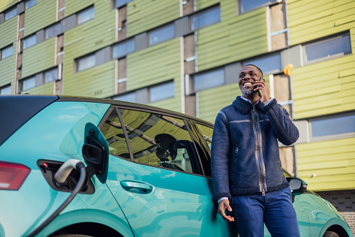 A low angle three quarter length shot of a mid adult businessman wearing formal businesswear taking a phone call on his smartphone while his electric car is on charge at a public charging point in Newcastle upon Tyne in the North East of England.