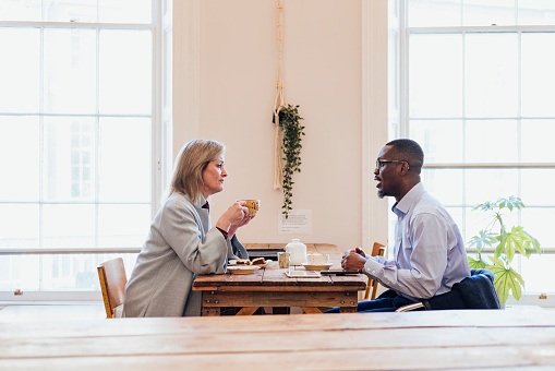 A side view wide shot of a mid adult businessman and a mature businesswoman wearing formal businesswear sitting at a table in a coffee shop talking business in the city, they are drinking hot drinks of tea and coffee, a teapot, smartphone and digital tablet are on the table in front of them.