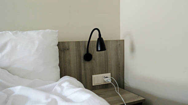 istock Close-up fragment of bedroom with empty bedside table, reading lamp and a USB socket in modern interior​ design home or hotel. Soft pillow and blanket, stylish comfortable furniture. Sun shadows. 1473462651
