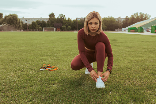 Serious athletic woman in sportswear tying shoelaces on sneakers during fitness training on sports field and looking at camera