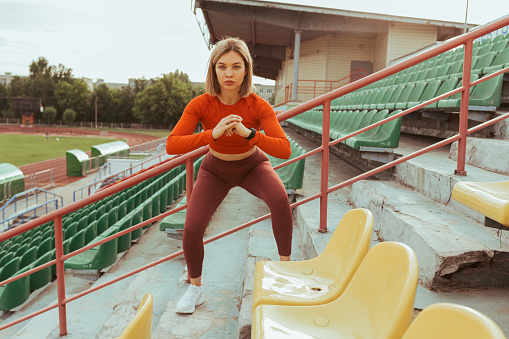 Confident athletic woman doing squat exercise and training near stadium seats in summer while looking at camera