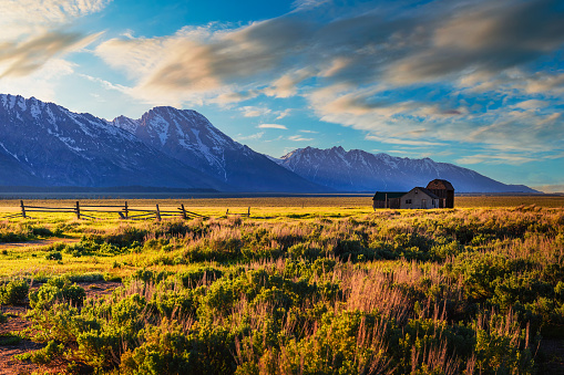Summer sunset over a historic farm house and the Thomas Murphy Barn at Mormon Row in Grand Teton National Park, with snowcapped Teton Mountain Range in the background.