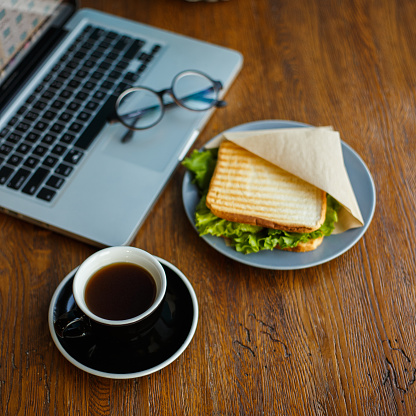 High angle of cup of delicious americano coffee arranged on wooden table with appetizing sandwich and laptop in cafe