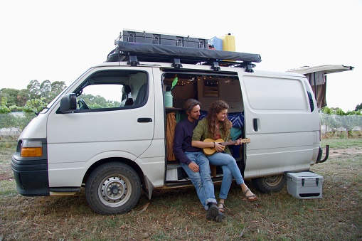 A young female and male traveler are sitting in a motor home. The woman is playing music on a ukulele.
