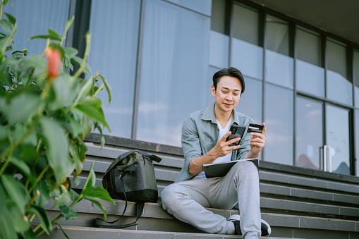 Young Asian man in a casual sitting on the stairs outside, holding a credit card and checking his bank account on his smartphone.