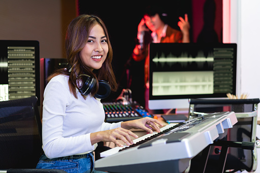 Asian producer woman in white shirt standing by sound mixing console. Happy female music composer artist with a man singer background