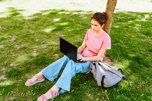 Student teenager, young woman, sitting on the grass in park, wearing jeans, pink t-shirt, with laptop, studying online, doing task, learning, social networking