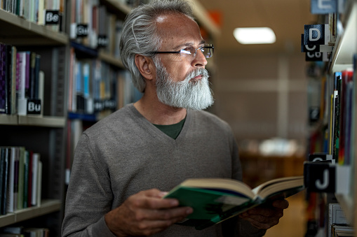 Waist-up side shot of a gray-haired retired man looking away while holding a book and standing between to big bookshelves at the city library during the afternoon.