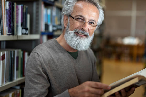istock Gray-Haired Man Smiling At Camera While Holding A Book At The Library 1473485194