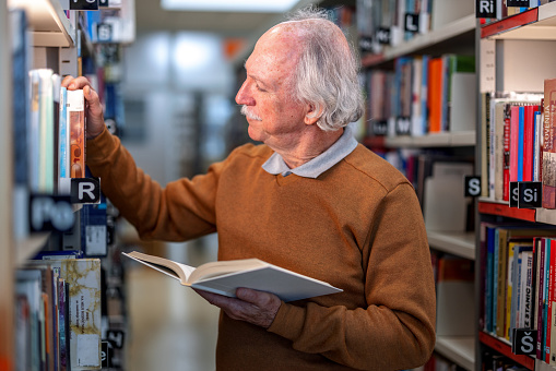 Waist-up side shot with blurred background of a formally dressed senior Caucasian man checking for book of his interest while standing between bookshelves and holding a book with white cover as a part of his weekend routine.