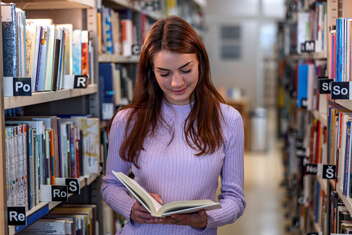 Waist-up front shot with blurred background of a young Caucasian university student reading a book as a preparation for her upcoming classes at the library.