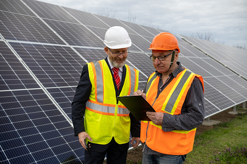 A bearded cheerful entrepreneur and a maintenance engineer are talking in the middle of a field of solar panels. They are wearing reflective vests and helmets.