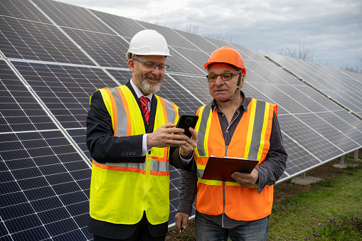 A bearded cheerful entrepreneur and a maintenance engineer are talking in the middle of a field of solar panels. They are wearing reflective vests and helmets. The entrepreneur shows the results to the engineer on his phone.
