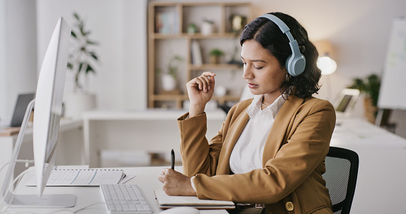 Headphones, computer and woman writing notes in office while streaming music, radio or podcast. Technology, business and female employee with notebook for internet research while listening to song.