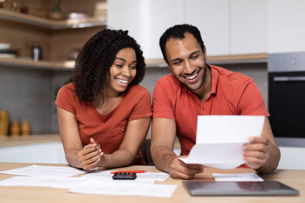 istock Happy young black lady in red t-shirt shows documents to guy with computer at kitchen interior 1473500842