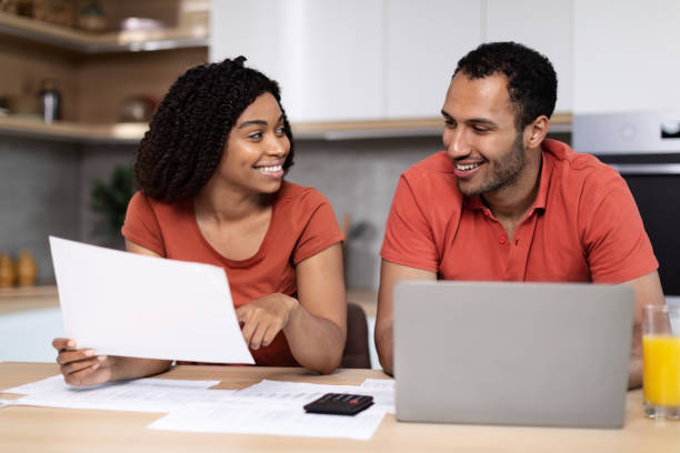 istock Smiling handsome young black woman in red t-shirt shows documents to husband with computer 1473500936