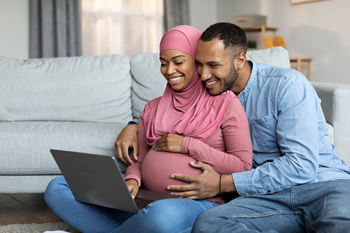 Loving Black Islamic Spouses Expecting Baby Using Laptop At Home, Happy Pregnant Muslim Couple Relaxing With Computer In Living Room, Shopping Online Or Buying Medical Insurance, Free Space