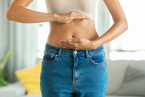 Unrecognizable Slim Woman Posing Framing Flat Stomach And Belly Button With Hands Standing Wearing Skinny Jeans At Home. Weight Loss And Abdomen Health Concept. Cropped