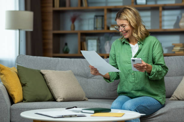 un'anziana donna sorridente siede a casa sul divano, tiene il telefono in mano, lavora con documenti, conti finanziari, affitto, prestiti, accordi - retirement senior adult planning finance foto e immagini stock