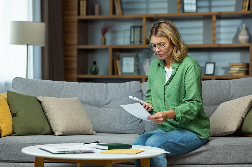 Serious and confused senior woman in glasses sitting on sofa at home. Worriedly holding the phone and documents in his hands. Problem with accounts, credit, payment.