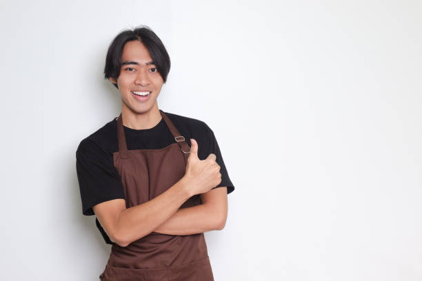 Portrait of attractive Asian barista man in brown apron with thumb pointing away on empty space. Advertising concept. Isolated image on white background stock photo