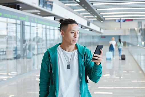 Young man wearing green hoodie using mobile phone while standing at airport.