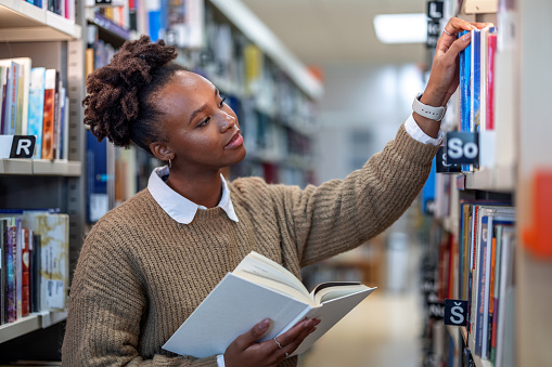 Waist up side shot with blurred background of a casually dressed young African-American female student smiling looking for a book at the city public library, surrounded with shelves full of books.