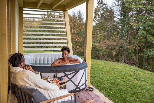 Diverse young adult Hispanic couple enjoying some time off outdoors at weekend getaway. He is sitting on a comfortable chair and she is enjoying in a hot tube outdoors.
