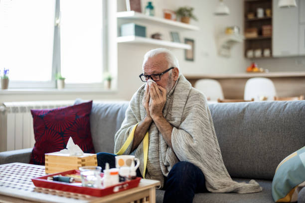 istock Senior man suffering from flu while sitting wrapped in a blanket on the sofa at home. Elderly Man blowing his nose while lying sick in bed at home. Man with a cold lying in sofa holding tissues. 1473504890