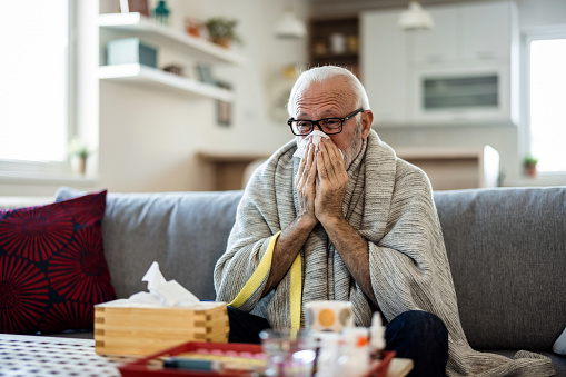 Cold exhausted senior man with flu wrapped in a warm blanket blowing his nose with a tissue in the livingroom. Man with a cold lying in sofa holding tissues.