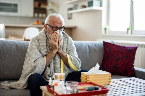 istock Senior man suffering from flu while sitting wrapped in a blanket on the sofa at home. Elderly Man blowing his nose while lying sick in bed at home. Man with a cold lying in sofa holding tissues. 1473505468